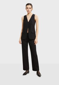 Černá vesta s pinstripem a hlubokým V-výstřihem, spárovaná s odpovídajícími upravenými kalhotami. Oba kousky mají jemné metalické akcenty a kapsy.