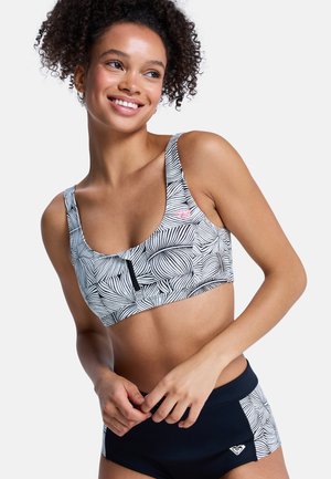 Czarny i biały wzorzysty sportowy biustonosz z zamkiem błyskawicznym z przodu, szerokimi ramiączkami i logo; w połączeniu z jednolitymi czarnymi szortami kąpielowymi.