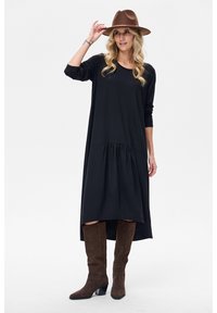 Czarna sukienka z długim rękawem, z marszczoną talią i asymetrycznym dołem, wykonana z miękkiego materiału. Noszona z brązowymi zamszowymi kozakami za kolano i brązowym kapeluszem.