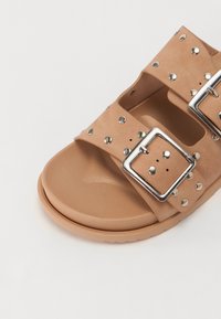 Infradito tan con due ampie cinghie con borchie argentate e fibbie rettangolari, suola piatta con una texture liscia.
