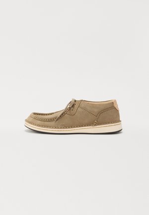 Scarpa in suede marrone con punta tonda, dettagli cuciti, suola beige e design con lacci. Parte superiore liscia con un leggero tacco e dettaglio sul retro.