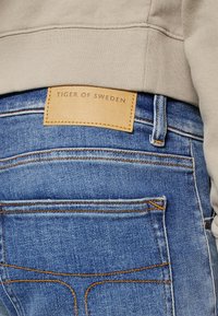 Blå denimjeans med en beige läderetikett som säger "TIGER OF SWEDEN." Har orange för stitching och en slät yta med bakfickor synliga.