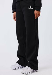Zwarte sweatpants van zacht materiaal, met zijzakken en een gestileerd wit logo op de linker dij, gecombineerd met dikke witte sneakers.