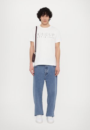 Hvid T-shirt med "Dondup Made in Italy" trykt i grå, kombineret med brede blå denimjeans. Modellen holder en lilla brun crossbody taske.