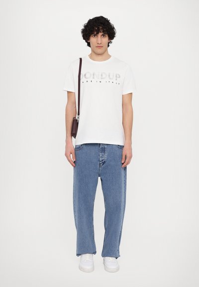 T-shirt blanc avec "Dondup Made in Italy" imprimé en gris, associé à un large jean en denim bleu. Le modèle tient un petit sac à bandoulière marron.