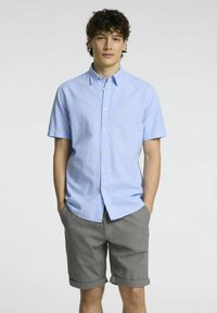 Camicia azzurra chiara a maniche corte con bottone in tessuto a trama semplice, abbinata a pantaloni grigi. I pantaloni sono arrotolati all'orlo, evidenziando un look informale.