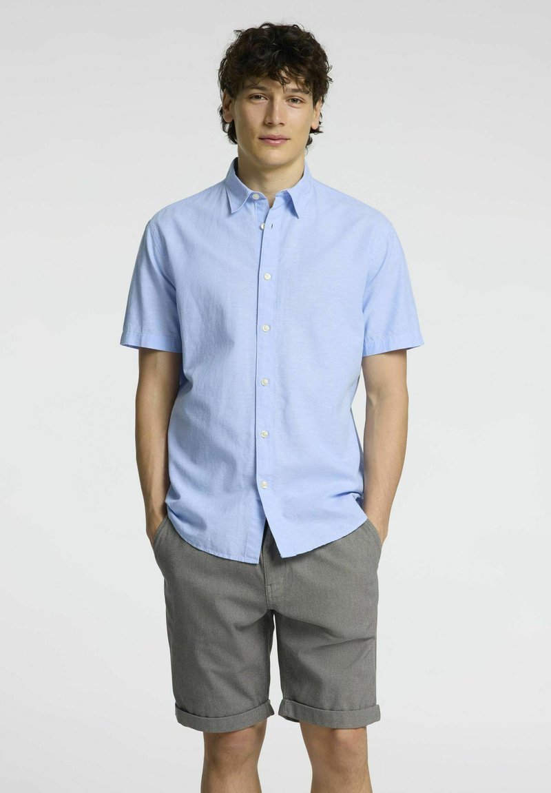 Camicia azzurra chiara a maniche corte con bottone in tessuto a trama semplice, abbinata a pantaloni grigi. I pantaloni sono arrotolati all'orlo, evidenziando un look informale.