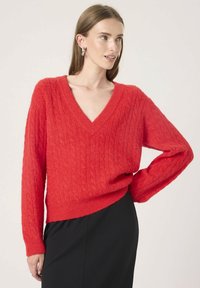 Woman wearing bright red cable-knit V-neck sweater and black skirt, posing with one hand on hip against plain background.