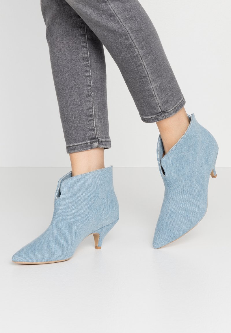 Personne portant un jean slim gris et des bottines bleu clair à bout pointu avec de petits talons sur un sol blanc.