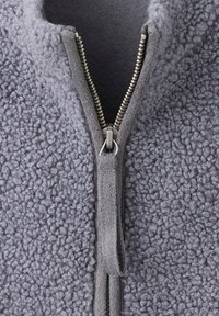 Gros plan d'une veste polaire grise avec une fermeture éclair argentée partiellement fermée et une languette en tissu sur le curseur de la fermeture éclair.