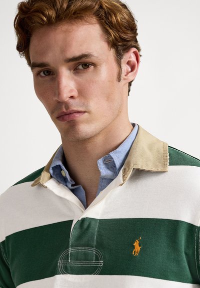 Jauns vīrietis ar gaisvas brūnas krāsas matiem, valkājot zaļu un baltu svītru regbija krekli ar gaiši brūnu apkakli virs gaiši zila krekla ar pogām.