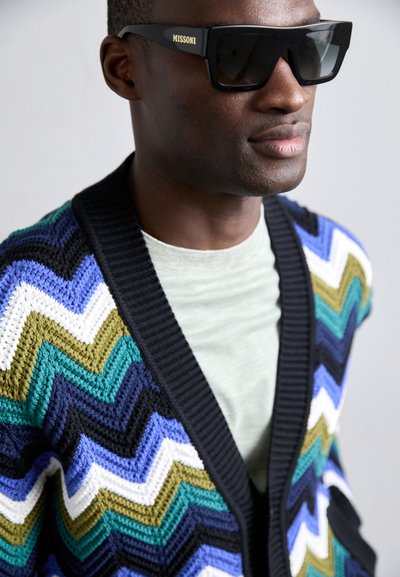 Cardigan croșetat în model zigzag cu culorile negru, albastru, verde și alb; echipat cu un guler ribbed negru; asortat cu ochelari de soare întunecați.