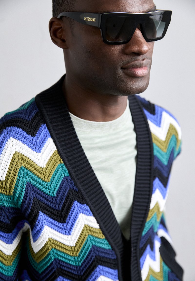 Cardigan all'uncinetto con motivo a zigzag nei colori nero, blu, verde e bianco; dotato di un colletto a coste nero; abbinato a occhiali da sole scuri.