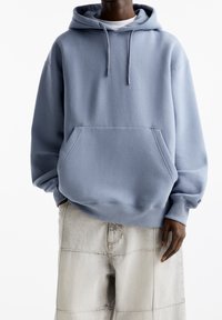 Persona con una sudadera holgada de color azul claro con bolsillo frontal y pantalones de pierna ancha beige frente a un fondo blanco.