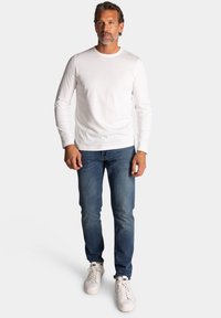 Witte lange mouwen overhemd, blauwe spijkerbroek en witte sneakers. Het overhemd heeft een ronde halslijn; de spijkerbroek is slim fit met een klassieke denim textuur.