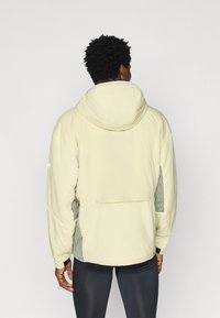 Světlě žlutá bunda s kapucí, která má texturované šedé boční panely a patentní knoflík. Hladká tkanina s jemně vyztuženým designem.