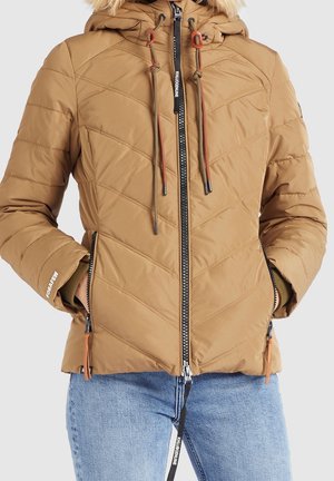 Veste matelassée beige avec capuche bordée de fourrure, fermeture éclair à l'avant et poches zippées, portée avec un jean bleu clair, mains dans les poches.