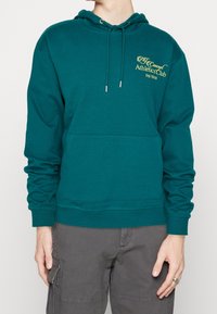 Osoba ubrana w turkusowy hoodie z przednią kieszenią i żółtym napisem "Concept Athletics Club (ny) 10019", w połączeniu z szarymi spodniami cargo.