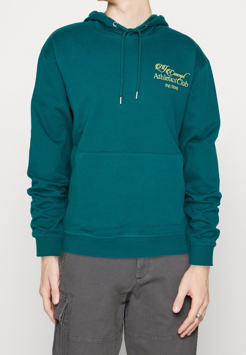 Osoba ubrana w turkusowy hoodie z przednią kieszenią i żółtym napisem "Concept Athletics Club (ny) 10019", w połączeniu z szarymi spodniami cargo.