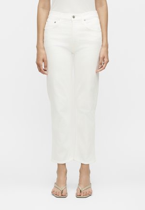 Γυναίκα που φοράει λευκά τζιν με ψηλή μέση και ίσια μπατζάκια και μπεζ πέδιλα με τακούνια, στέκεται μπροστά σε λευκό, απλό φόντο.