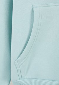 Šviesiai mėlynos spalvos džemperio audinys su tekstūruota paviršiaus struktūra ir priekinė kišenė. Apatinė kraštinė ir kišenės kraštai siūti tampria juosta, siekiant didesnio patvarumo.