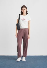 Νεαρή γυναίκα που στέκεται φορώντας ένα λευκό κοντομάνικο μπλουζάκι Nike, μωβ παντελόνια σε ίσια γραμμή, λευκά αθλητικά παπούτσια και μια μαύρη τσάντα χιαστί σε έναν ουδέτερο φόντο.