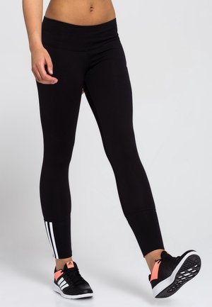 Zwarte leggings met een hoge tailleband, gemaakt van een gladde, rekbare stof en met witte strepen langs de onderkant van de buitenzijde van het been. Gepaarde met zwarte sneakers.