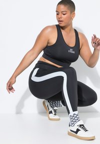 Top crop atletico nero con la scritta "GET STRONG", abbinato a leggings neri a vita alta con una striscia laterale bianca. Gli accessori includono calze a fantasia e sneakers.