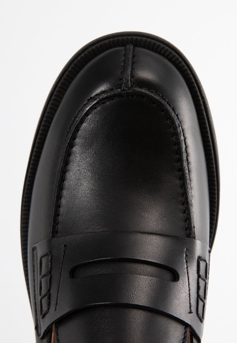 Nahaufnahme eines schwarzen Lederloafer mit detaillierter Naht und einem Riemen über dem oberen Teil, auf einem weißen Hintergrund.