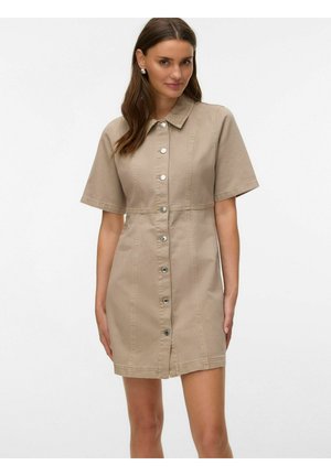 Vrouw die een beige kortmouwig jurk met knopen en kraag draagt, staand tegen een effen lichte achtergrond.