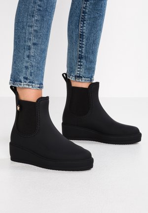 Person iført sorte ankelstøvler med elastiske sidepaneler og trækketab, kombineret med blå denimjeans mod en hvid baggrund.