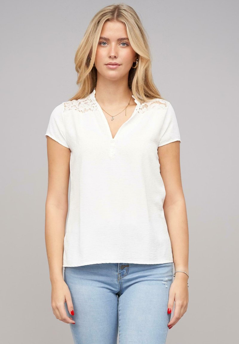 Haut blanc à manches courtes avec un accent en dentelle sur les épaules, présentant un col en V et une patte de boutonnage ; tissu lisse et léger.