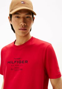 Rød bomulds T-shirt med korte ærmer, som har et sort logo med teksten "TOMMY HILFIGER" og yderligere tekst. Beige kasket med broderet logo.