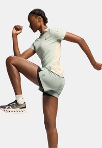 Ljusgrön träningsskjorta och shorts med svart foder, med en strömlinjeformad design och en logotyp på shortsen. Modell i en aktiv pose.