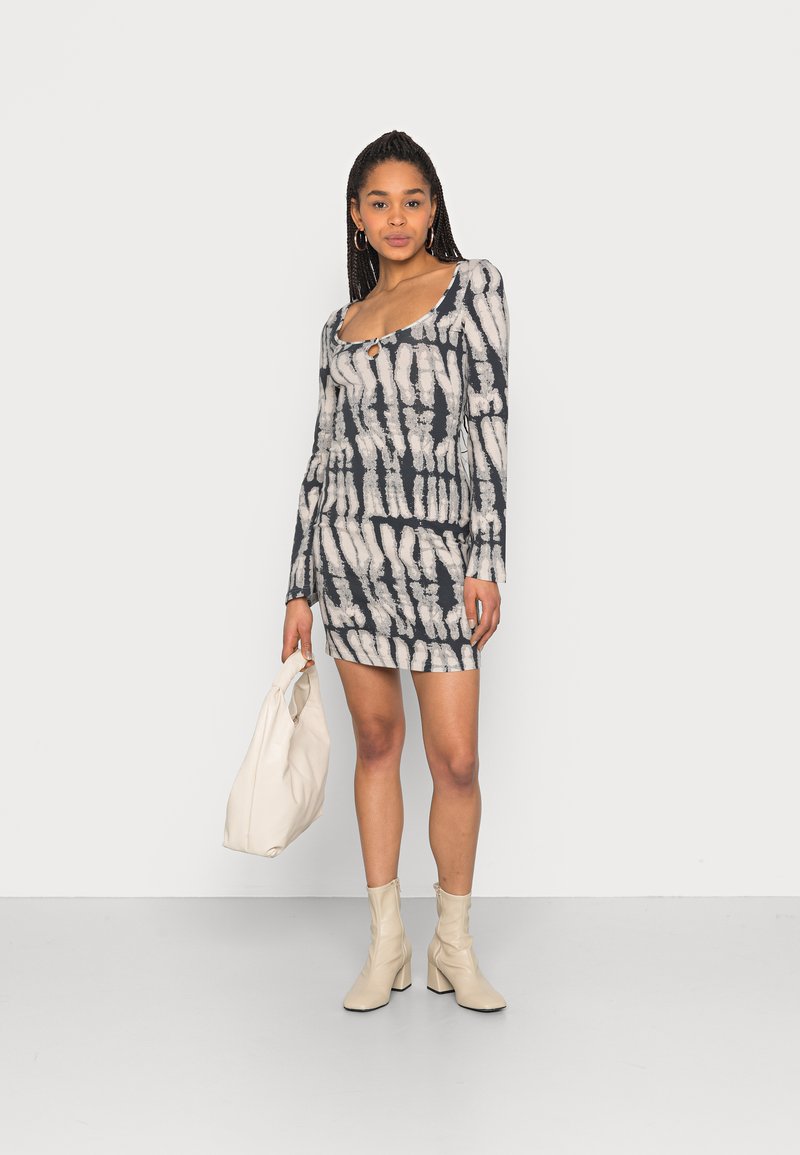 Vestido justo com mangas longas, padrão tie-dye em preto e creme, decote redondo e detalhe em fenda. Combinado com uma mala bege e botas de salto.