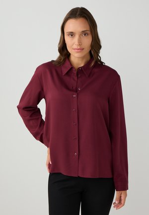 Vrouw in een burgundy blouse met lange mouwen en zwarte broek, staand met één hand op haar heup tegen een lichtgrijze achtergrond.