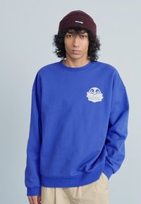Sweatshirt azul com gola redonda e mangas longas. Apresenta um logo branco no lado esquerdo do peito. Tecido macio com um ajuste descontraído.