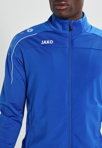 Blauwe sportjas van gestructureerde stof, met een hoge kraag, volledige ritssluiting, witte bies en het 'JAKO' logo op de borst.