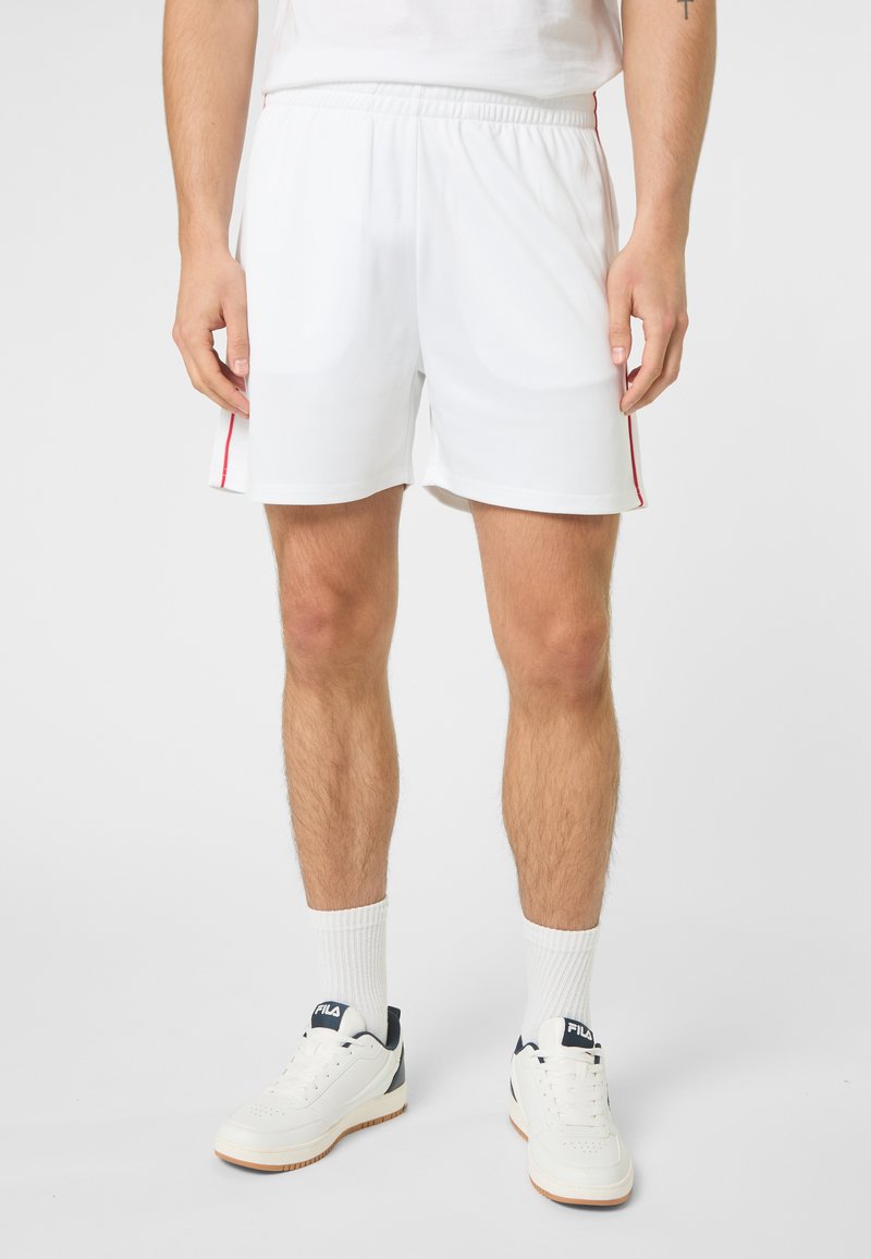 Homme portant un short de sport blanc avec des bandes rouges sur les côtés, des chaussettes blanches montantes et des baskets FILA blanches, debout devant un fond uni.