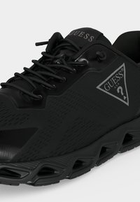 Baskets noires Guess avec logo triangulaire sur le côté, design à lacets et semelle sculptée avec des détails découpés.