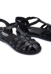 Sandalias de cuero negro con correas trenzadas, suela plana, correa ajustable en el tobillo y un acabado suave. Diseñadas para comodidad y transpirabilidad.