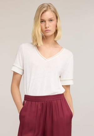 Giovane donna con capelli biondi fino alle spalle, che indossa una maglietta bianca con scollo a V e pantaloni bordeaux con vita elasticizzata, mani nelle tasche.