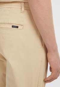 Beige shorts met een achterzak en O'Neill-logo label, gedragen door een persoon met een zichtbare rechterhand en onderarm.
