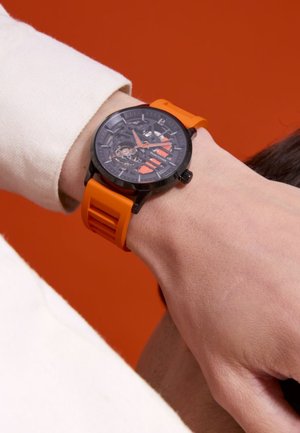 Zwart skelet horloge met oranje accenten en een siliconen band. De band heeft uitgesneden details en de wijzerplaat toont een ingewikkelde mechanische beweging.