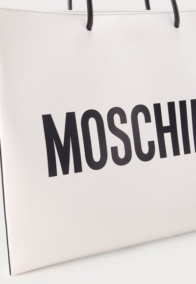 Sac cabas blanc avec des poignées noires affichant un grand texte noir « MOSCHINO » partiellement visible sur le devant.
