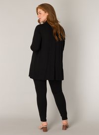 Zwarte langslavige top met een ontspannen pasvorm, voorzien van een gebogen onderrand, gecombineerd met zwarte leggings en sandalen met blokhakken en bandjes.