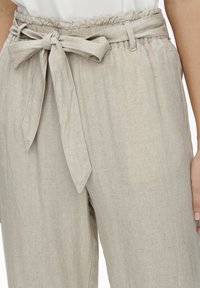Pantalons en lin beige clair avec une taille élastique, des poches latérales et une ceinture nouée. Le tissu a une finition légèrement texturée et respirante.