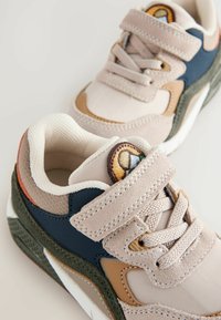 Sneakers with beige, navy, and green accents. Mesh and suede materials. Features velcro strap and laces. Round logo patch on the tongue.