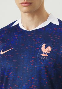 Maglietta da soccer con motivi blu e rossi, colletto bianco, con un emblema di gallo e il logo Nike arancione nella zona del petto.