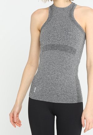 Kvinde iført en ærmeløs, grå, tætsiddende træningstop og sorte leggings, stående foran en ensfarvet hvid baggrund.